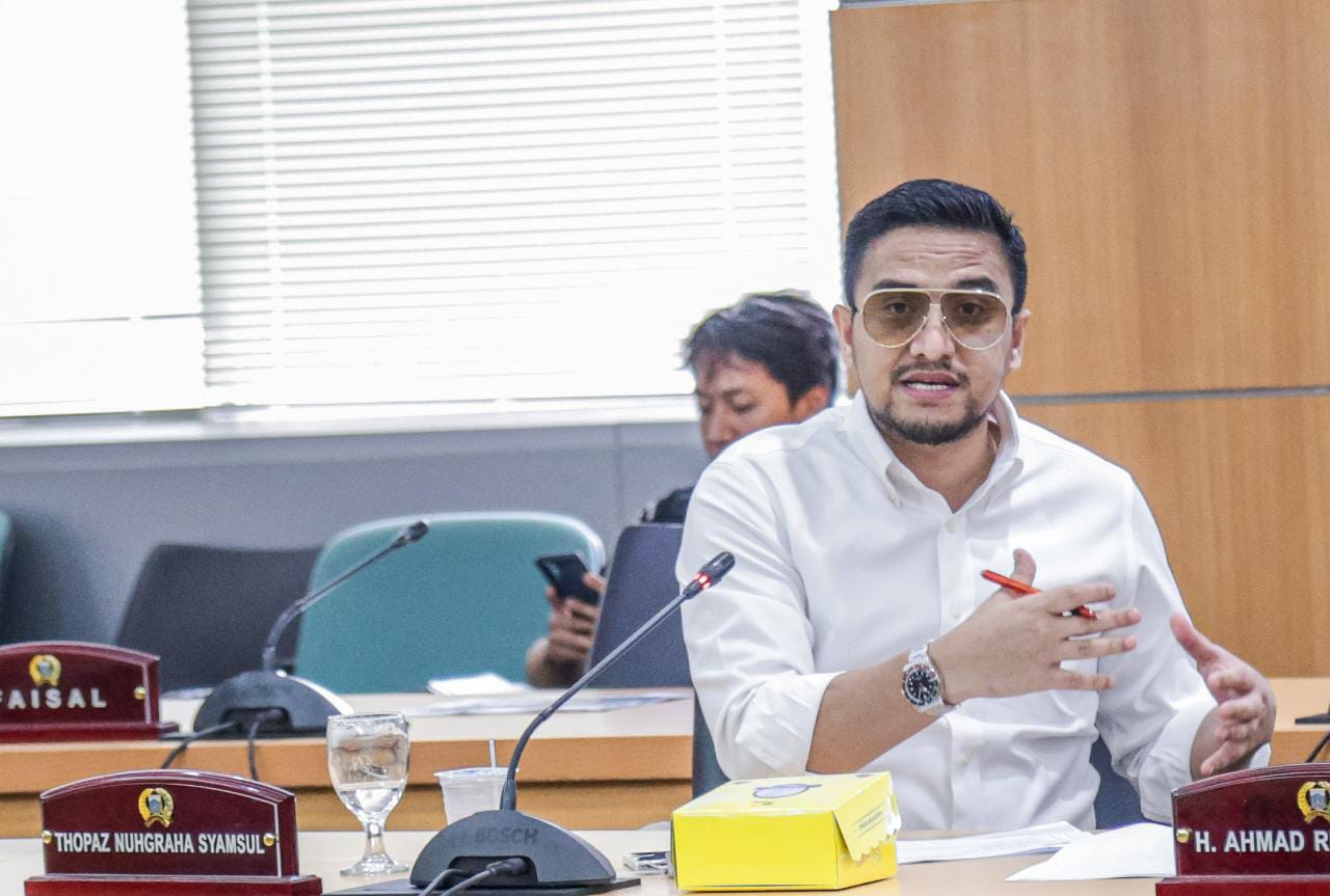 Thopaz mengenakan kemeja putih dan berbicara melalui mikrofon di ruang rapat.